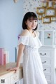 A woman in a white dress standing next to a dresser.