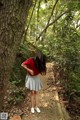 A woman in a red shirt and gray skirt standing on a path in the woods.