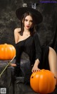 A woman in a black dress and hat sitting next to a pumpkin.