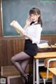 A woman sitting at a desk in front of a blackboard holding a book.