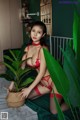 A woman in a red lingerie sitting on a bed next to a plant.