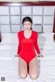 A woman in a red bodysuit sitting on a bed.