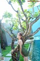 A woman in a green dress leaning against a tree by a pool.